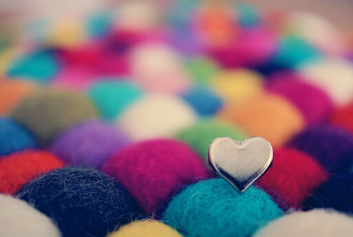 Diverse Wollknäuel in beige, dunkelblau, hellblau, gelb, pink, lila und rot. Auf einem Knäuel steckt ein silbernes Herz.